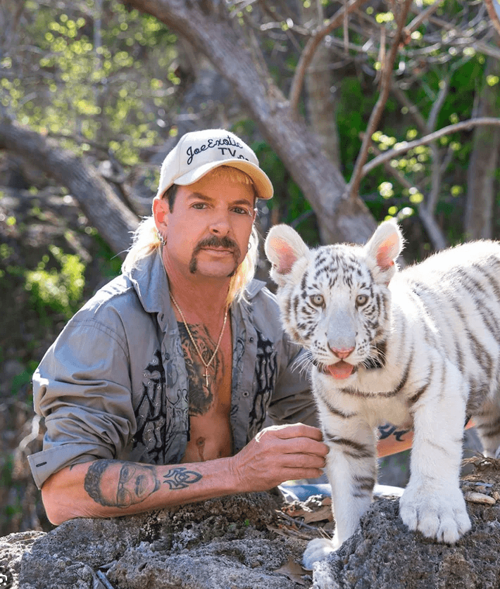 Man in a Joe Exotic TV cap posing beside a white tiger cub on a rock ledge.